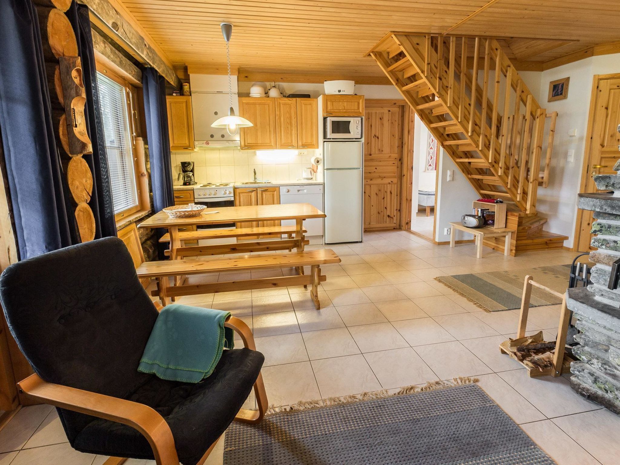 Een gezellig houten interieur met een eettafel, een bankje, een stoel met een plaid, een keuken met houten kasten, een magnetron, een fornuis en een stenen trap.