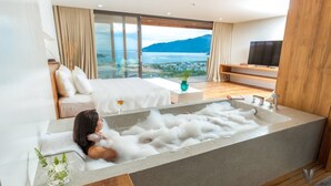 Een moderne badkamer met een groot bad vol met bubbels, een glas wijn en uitzicht op een bergachtig landschap.