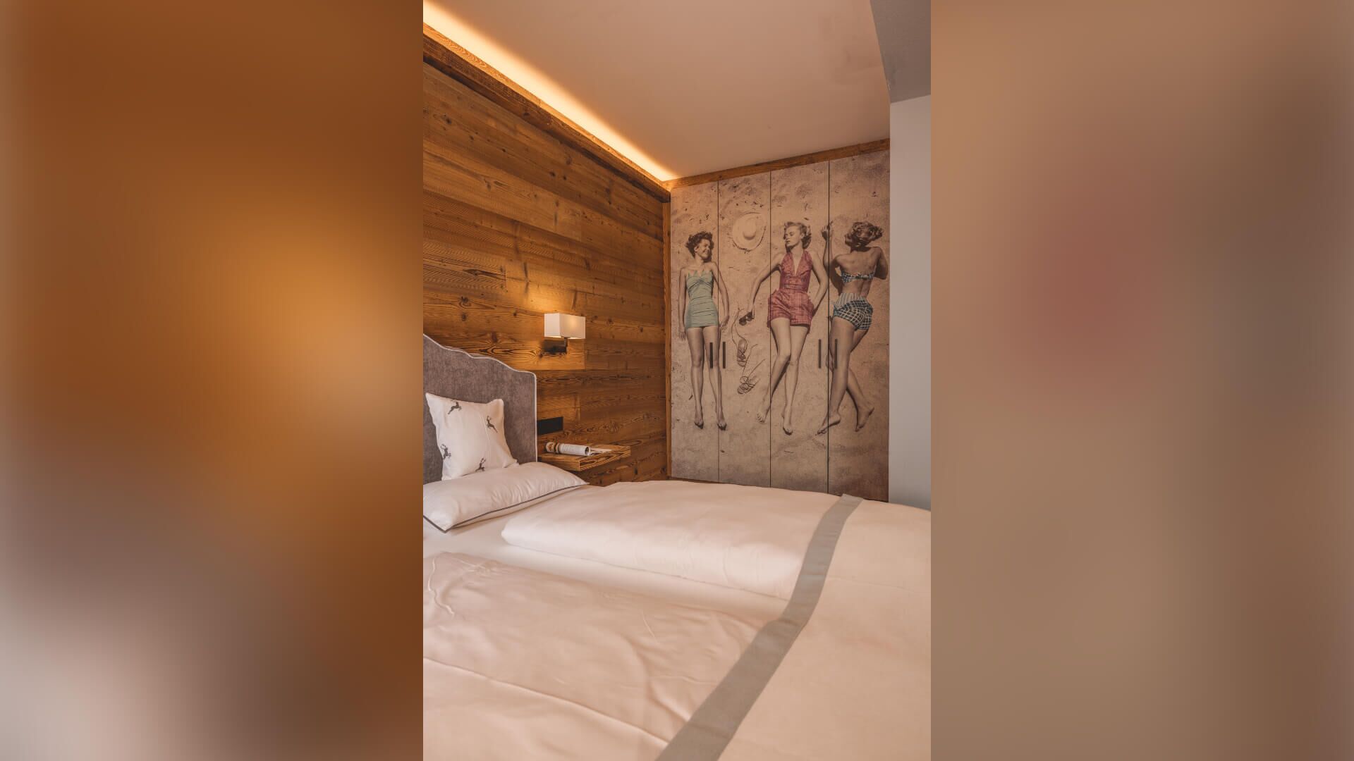 A bedroom with a wooden headboard, a bed with white linens, a nightstand with a lamp, and a wall mural of two figures.
