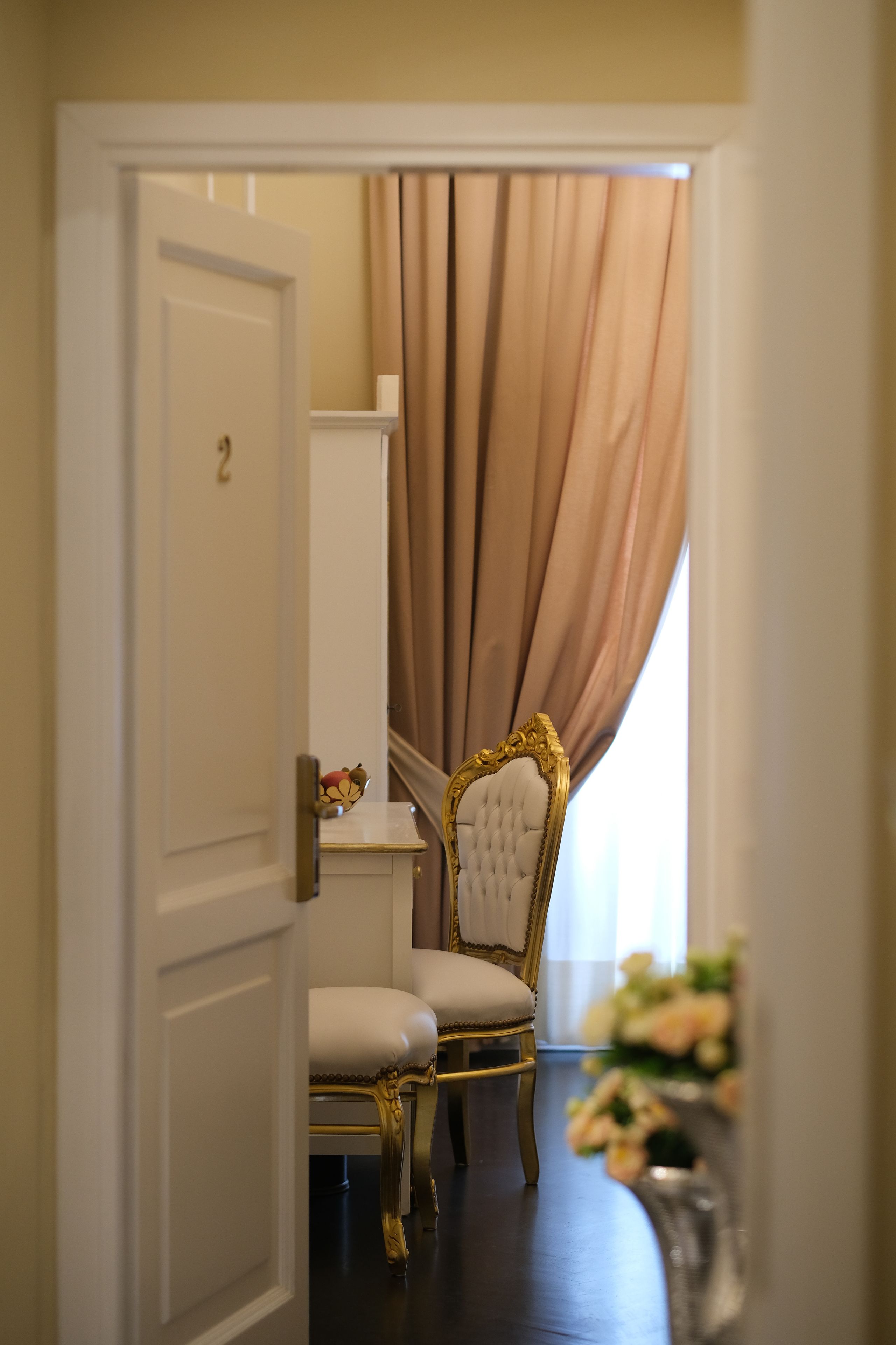 A room with a white door, a chair with gold detailing, a white table, and a curtain.