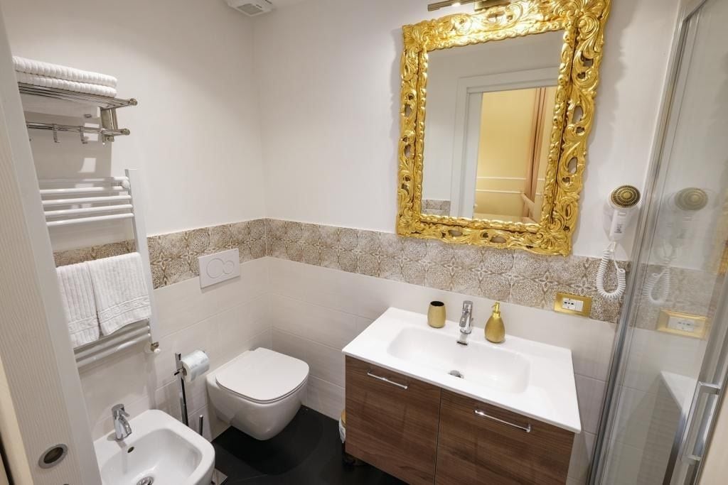 Ein Badezimmer mit einem weißen Waschbecken, einem spiegel mit goldener Rahmung, einer weißen Toilette und einer Duschkabine aus Glas.