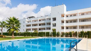 Una piscina con superficie de agua azul cristalina, rodeada por una terraza pavimentada. Al fondo, un edificio blanco de varios pisos con balcones y ventanas.