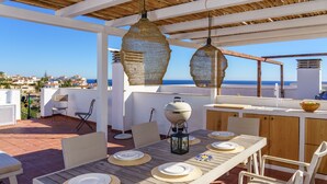 Un patio al aire libre con una mesa de madera dispuesta para comer, una parrilla y lámparas de mimbre colgantes.