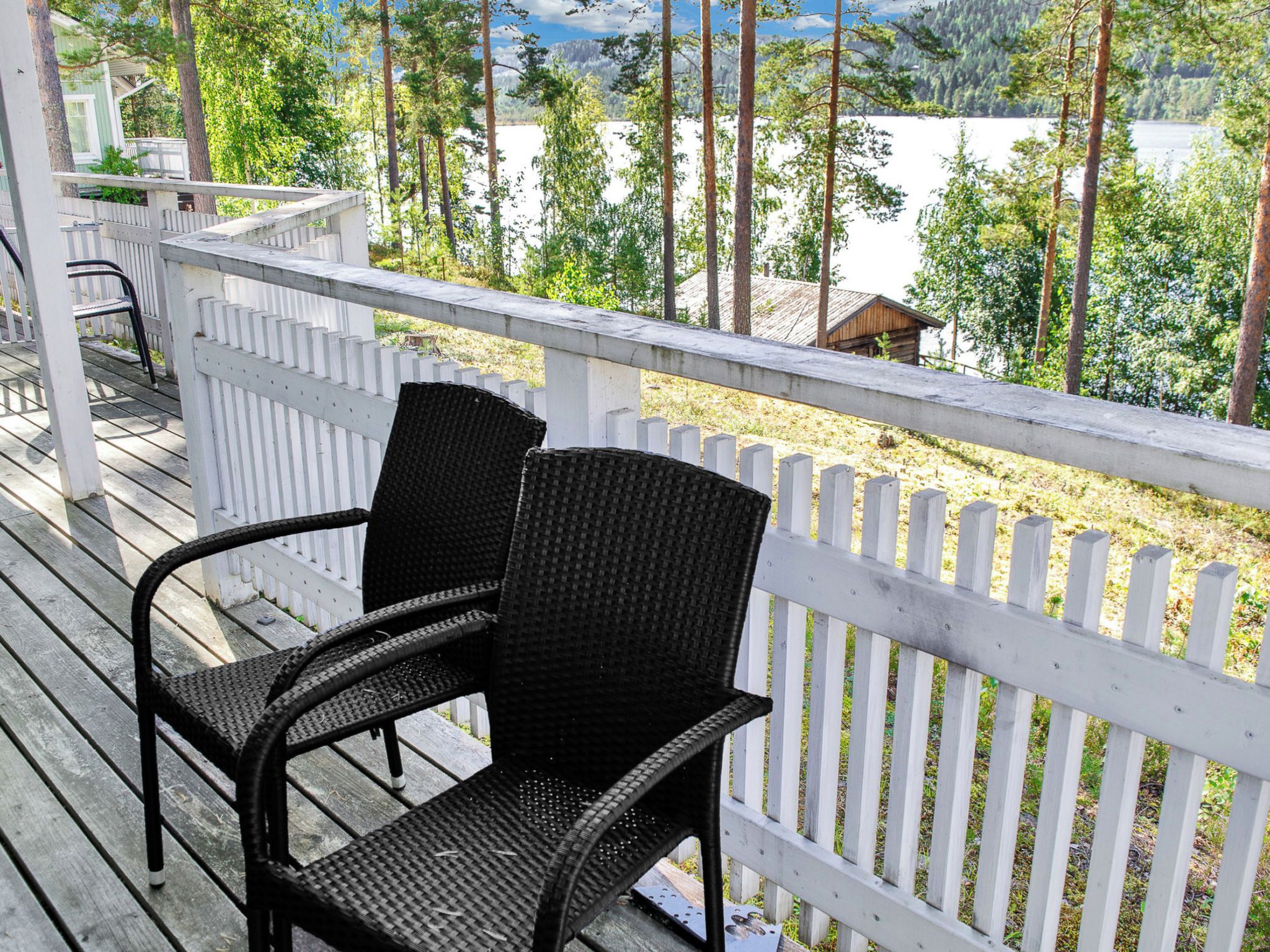 A wooden deck with two wicker chairs, a white picket fence, and a view of a lake and forest.