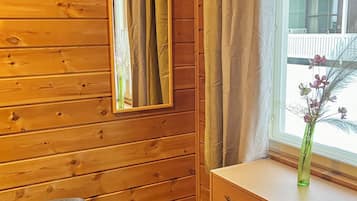 A wooden room with a mirror, a window with curtains, a bedside table with a vase, and a bed.
