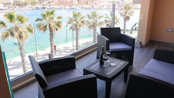 Un balcón con mobiliario de mimbre, una mesa con una botella y vasos, y vistas a un puerto deportivo con barcos y palmeras.