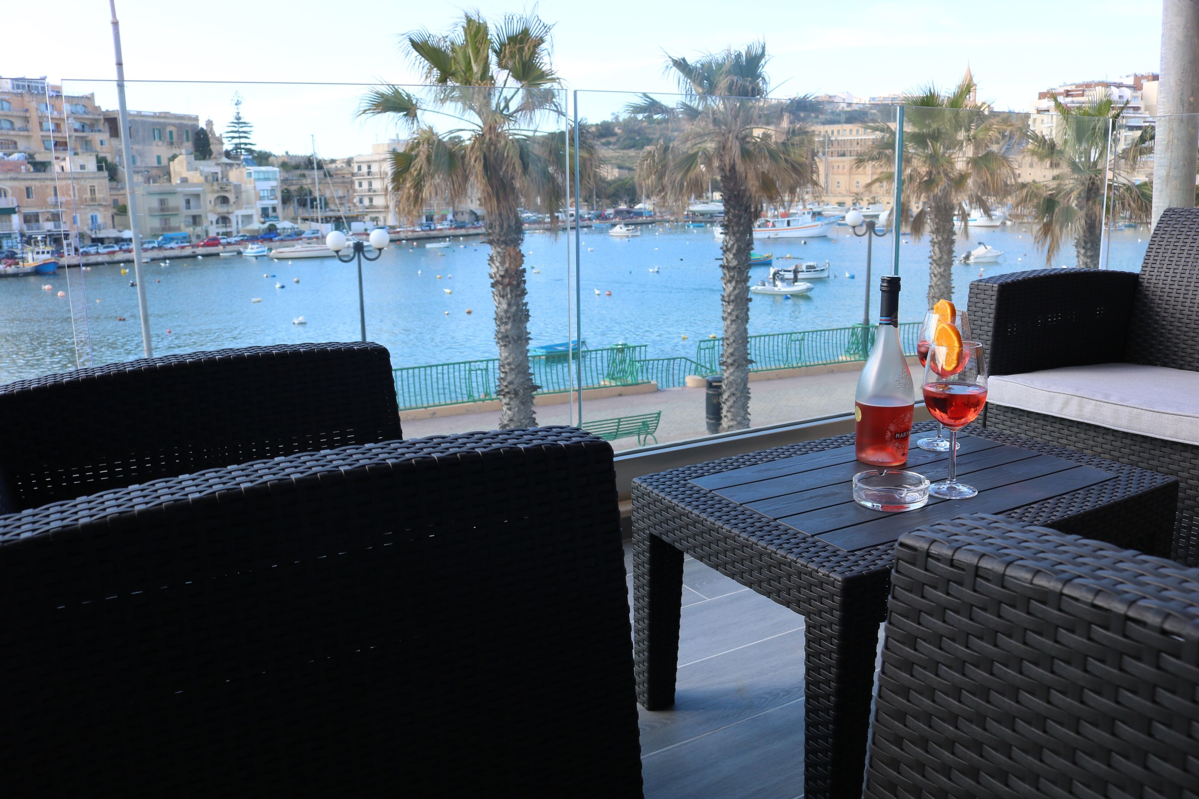 A balcony with wicker furniture overlooking a marina with boats and palm trees.