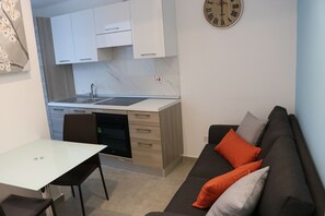 Une cuisine compacte avec des meubles blancs, un four encastré et un évier. On y trouve une table à manger avec des chaises, un canapé avec des coussins et une horloge murale.