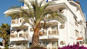 A multi-story hotel with balconies, a palm tree, and purple flowers in the foreground. - Villa Gizem (Manavgat)