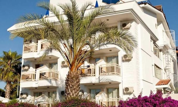 A multi-story hotel with balconies, a palm tree, and purple flowers in the foreground. - Villa Gizem (Manavgat)