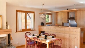 A cozy kitchen with wooden cabinets and a central island, a dining area with a table and chairs, and a stone fireplace. - Romarin 220 by Interhome (Verbier)