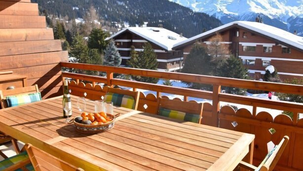 A wooden deck with a table set for dining, overlooking a mountainous landscape with snow-capped peaks and buildings in the background. - Romarin 220 by Interhome (Verbier)