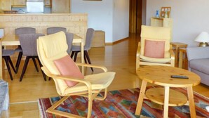 A spacious living room with a wooden dining table, two armchairs, a sofa, and a kitchen area with wooden cabinets and a hanging light fixture. - Romarin 220 by Interhome (Verbier)