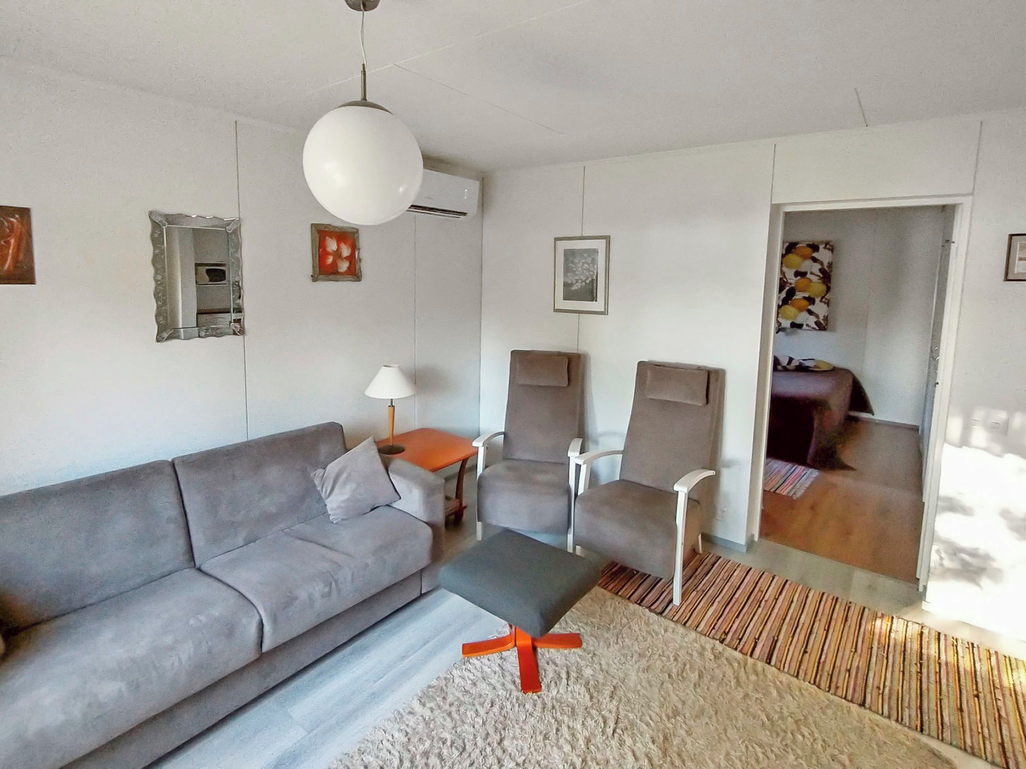 Un salon moderne avec un canapé gris, deux fauteuils, une petite table et une lampe. On y trouve également un ventilateur de plafond blanc, un tapis et un plancher en bois.