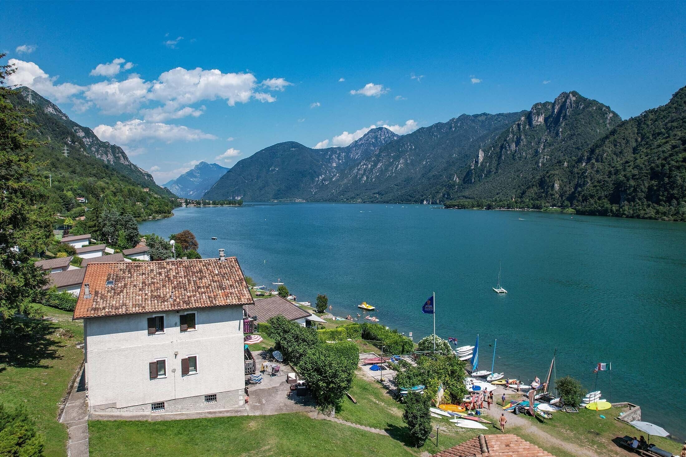 A lakeside property with a white building, a dock with boats, and mountains in the background.