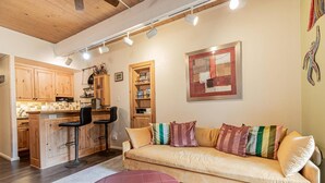 Una sala de estar acogedora con sofá, reposapiés y una cocina con armarios de madera y taburetes de bar.