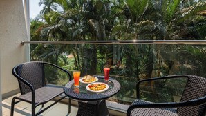 A balcony with a glass railing, two wicker chairs, a small round table, and a view of dense tropical foliage. - Regenta Central North Goa (Arpora)