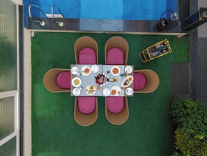 Un área de comedor al aire libre con una mesa dispuesta para cuatro personas, rodeada de sillas de mimbre con cojines y una piscina al fondo.