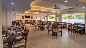 A restaurant with multiple dining tables and chairs, ceiling fans, and large windows with a view of greenery. - Regenta Central North Goa (Arpora)