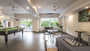 Ein geräumiger Innenbereich mit Billardtisch, einem Schachbrett und einem Essbereich mit Stühlen und Tisch. Deckenventilatoren, große Fenster und eine Tür, die nach draußen führt, sind vorhanden.