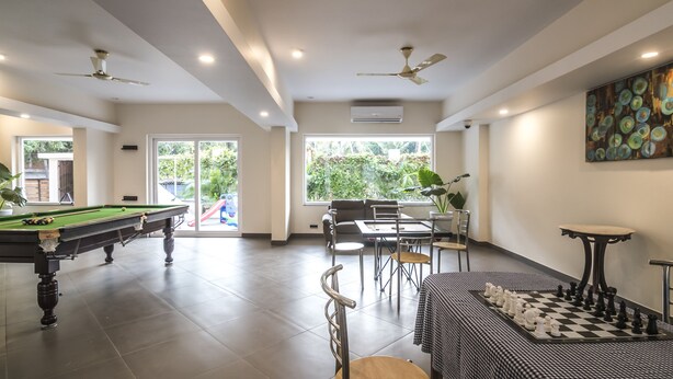 A spacious indoor area with a pool table, a chess table, and a dining area with chairs and a table. There are ceiling fans, large windows, and a door leading outside. - Regenta Central North Goa (Arpora)