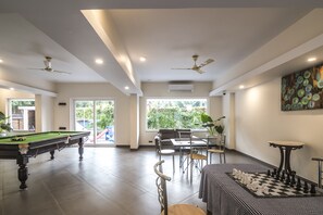 Amplio espacio interior con mesa de billar, mesa de ajedrez y comedor con sillas y mesa. Hay ventiladores de techo, ventanas grandes y una puerta que da al exterior.