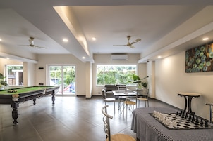 Amplio espacio interior con mesa de billar, mesa de ajedrez y comedor con sillas y mesa. Hay ventiladores de techo, ventanas grandes y una puerta que da al exterior.