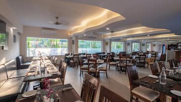 A spacious dining area with multiple tables and chairs, large windows offering a view of greenery, and a modern ceiling design.