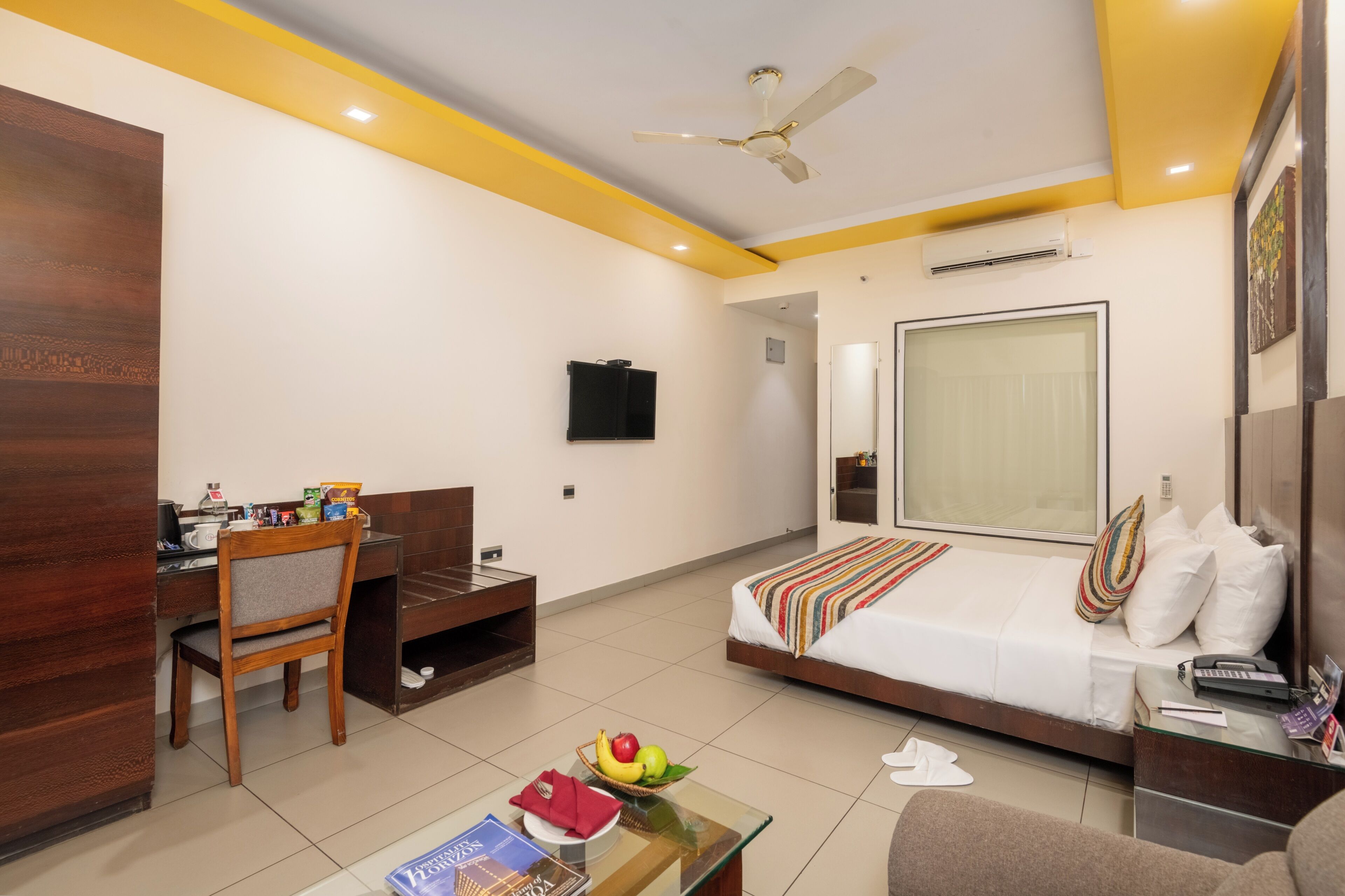 Ein Hotelzimmer mit Bett, Schreibtisch, Stuhl, Fernseher und Deckenventilator.