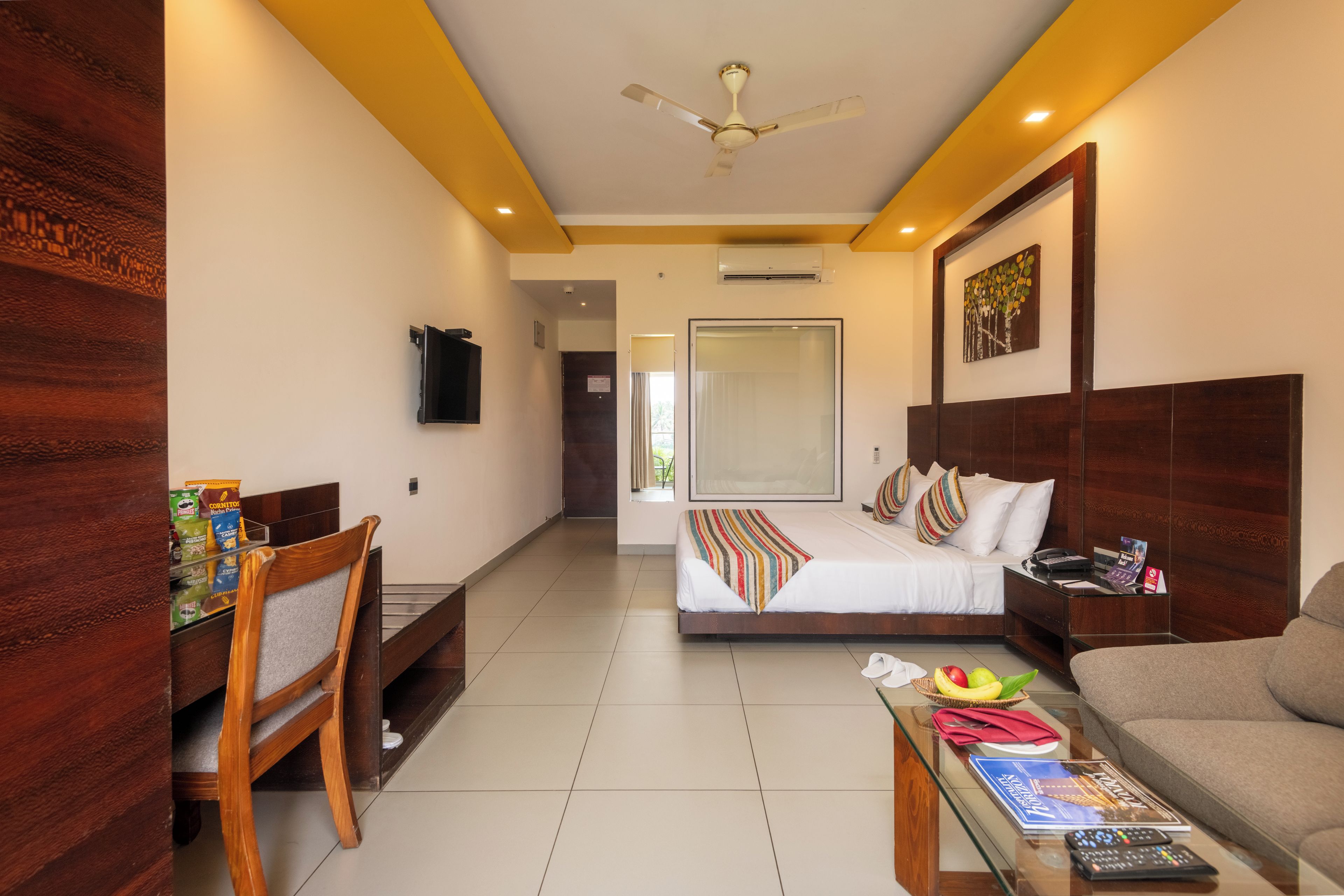 Ein Hotelzimmer mit Bett, Schreibtisch, Sessel, Sofa, Fernseher und Deckenventilator.
