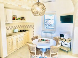 A kitchen with a round dining table, wicker chairs, a hanging light fixture, a window with a view, and a microwave on a small table.