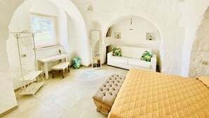 Une pièce avec un canapé blanc, un banc en bois, un lit et une petite table. Il y a deux fenêtres en arc de cercle et un miroir au mur.