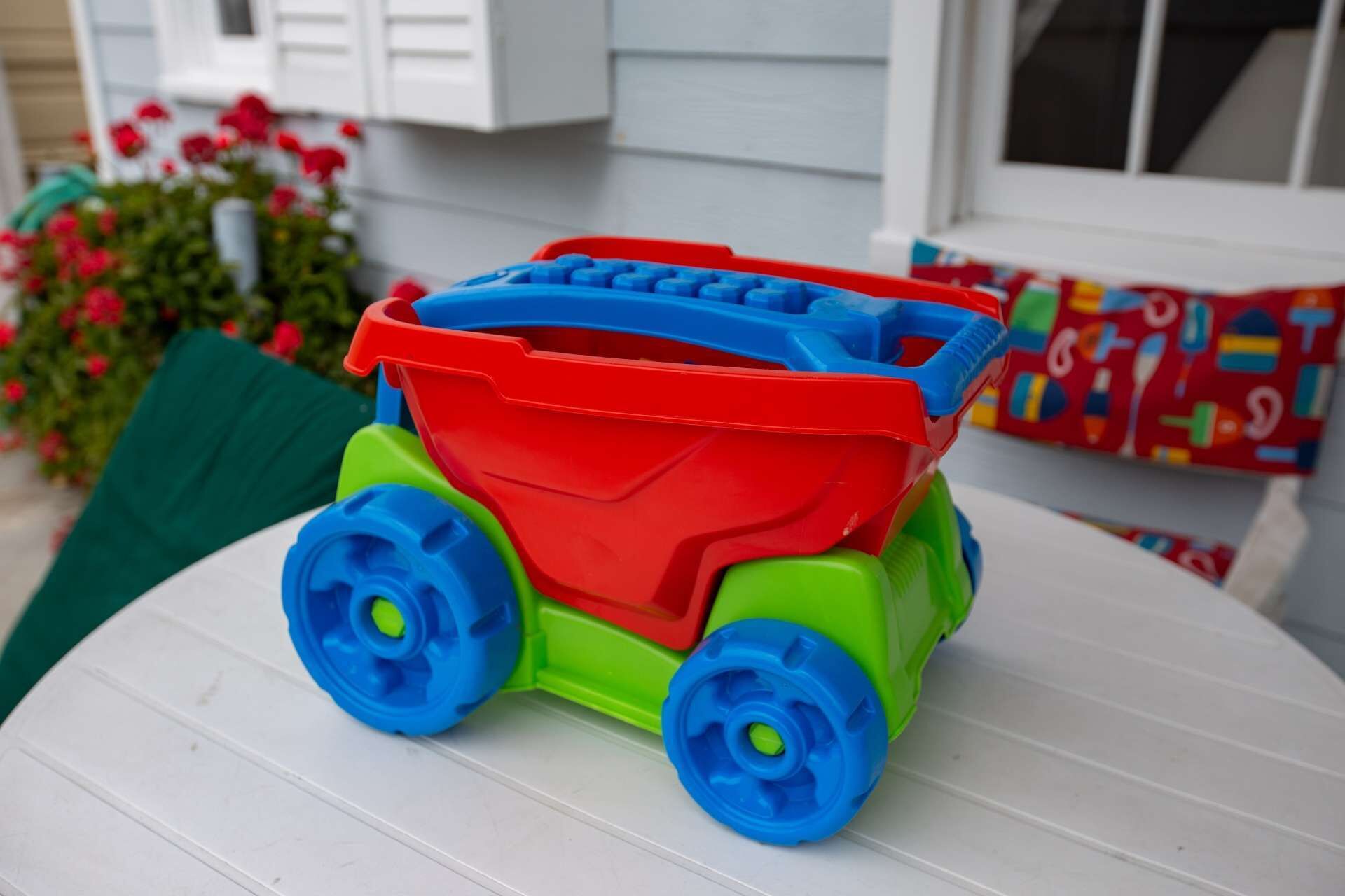 Um caminhão basculante de brinquedo sobre uma mesa branca, com uma casa e plantas ao fundo.