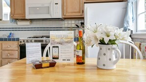 Eine Küche mit Holzausstattung, einer Mikrowelle, einem Herd und einem weißen Kühlschrank. Auf der Arbeitsplatte steht eine weiße Vase mit weißen Blumen, eine Weinflasche und eine Schachtel Pralinen.