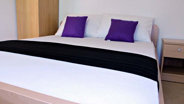 Een bed met een zwart-wit sprei, twee paarse kussens en een houten nachtkastje met een lade.