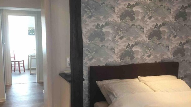 A bedroom with a wooden floor, a bed with white bedding, a dark headboard, and a patterned wallpaper. - Guest apartment on Petersberg with balcony (Erfurt)