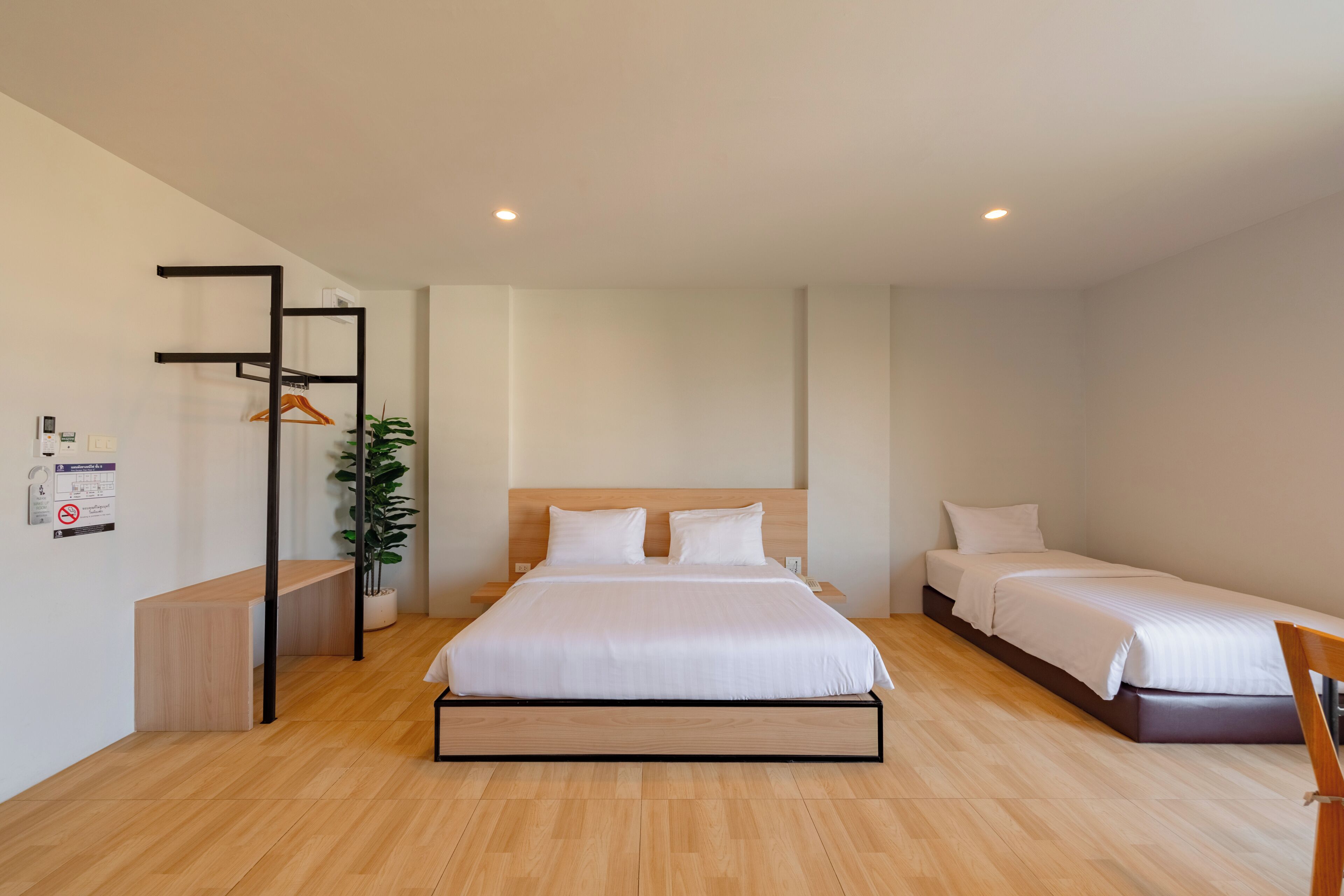 A modern hotel room with a large bed, a smaller bed, a desk, a plant, and a wall-mounted shelf.