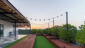 Un espacio al aire libre cubierto con piso a cuadros, bancos de madera, césped artificial e iluminado con guirnaldas de luces.