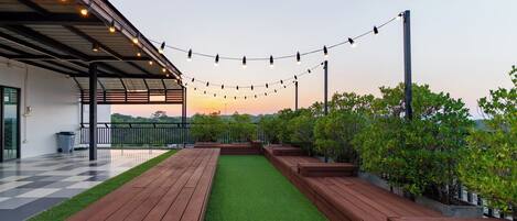 A covered outdoor area with a checkered floor, wooden benches, artificial grass, and string lights.