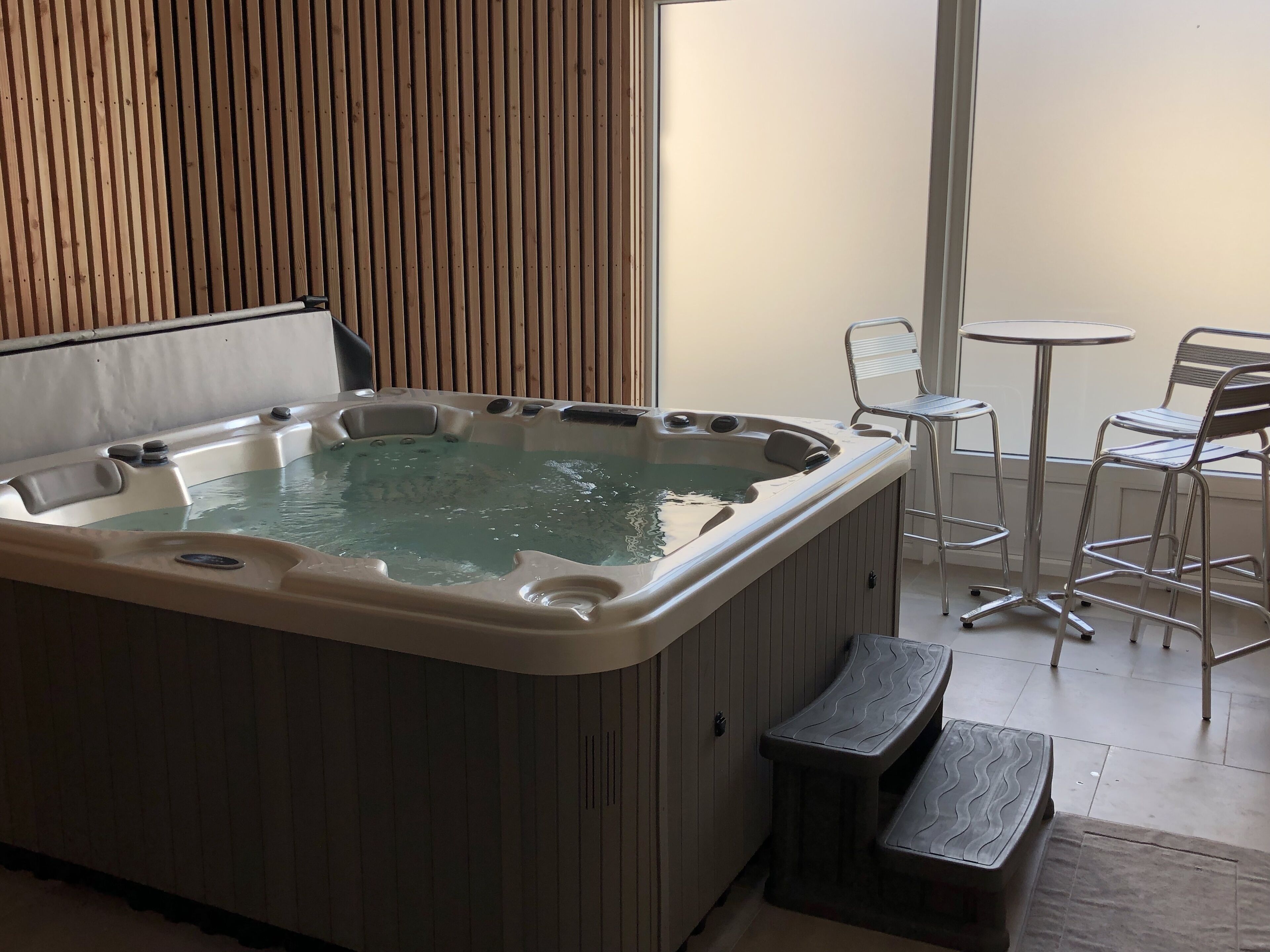 A hot tub with a wooden panel backdrop, a small table, and two chairs.