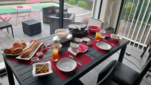 A dining table set with breakfast items, including croissants, fruit, and a hot plate, overlooking a backyard with a pool and outdoor furniture. - COSY BEDROOM 23m2. Adults Only (agde)