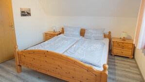 A wooden bed with a grey bedspread and pillows, two bedside tables with lamps, a wooden door, and a small picture on the wall. - Vacation home Nemo, holiday apartment rental North Sea - Holiday home Nemo (Varel)