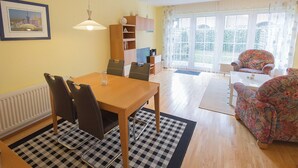 A dining area with a table and chairs, a bookshelf, and a window with curtains. - Vacation home Nemo, holiday apartment rental North Sea - Holiday home Nemo (Varel)