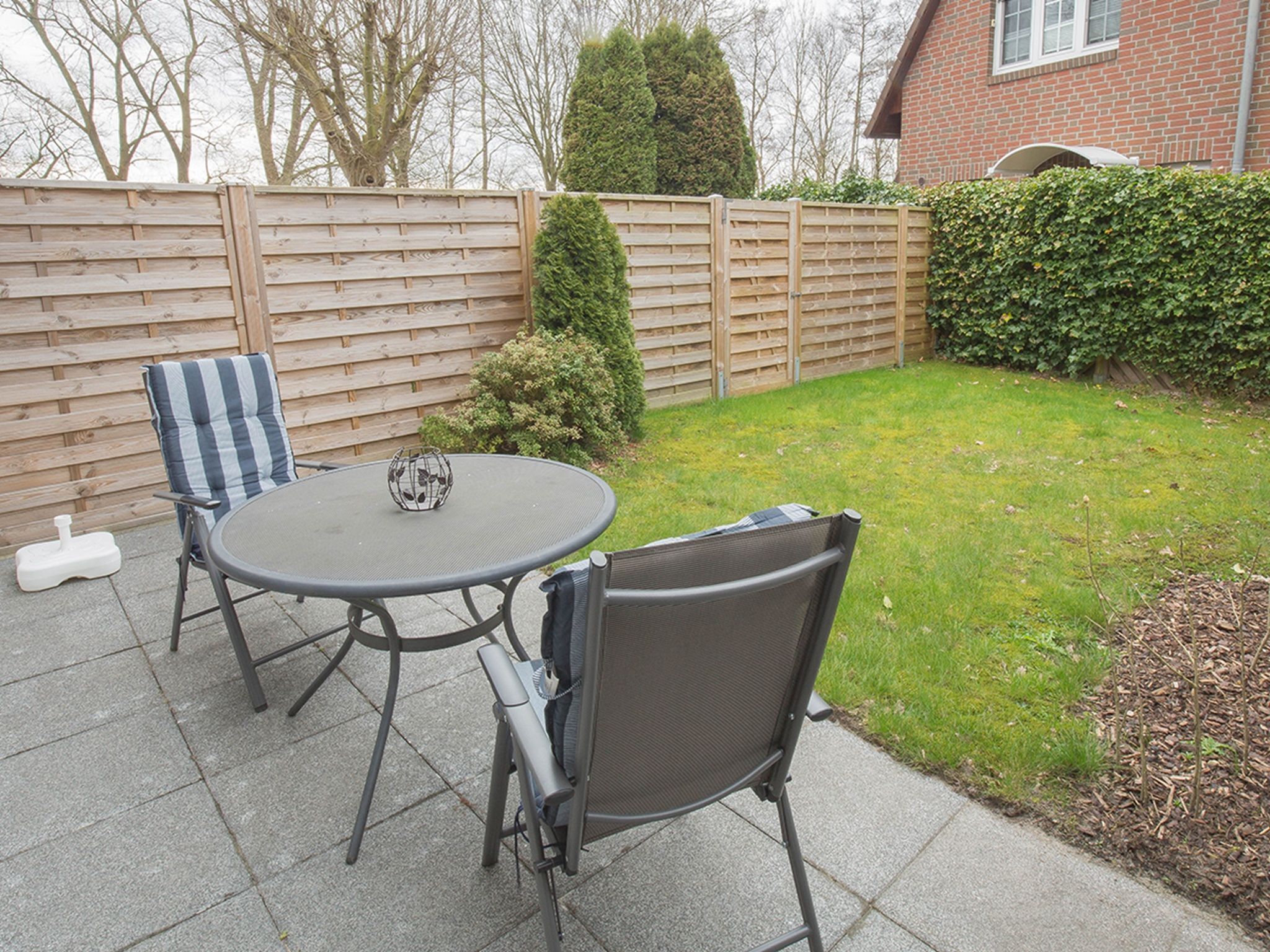 Een terras met een ronde tafel en twee stoelen, een klein decoratief voorwerp op de tafel, en een houten hek op de achtergrond.