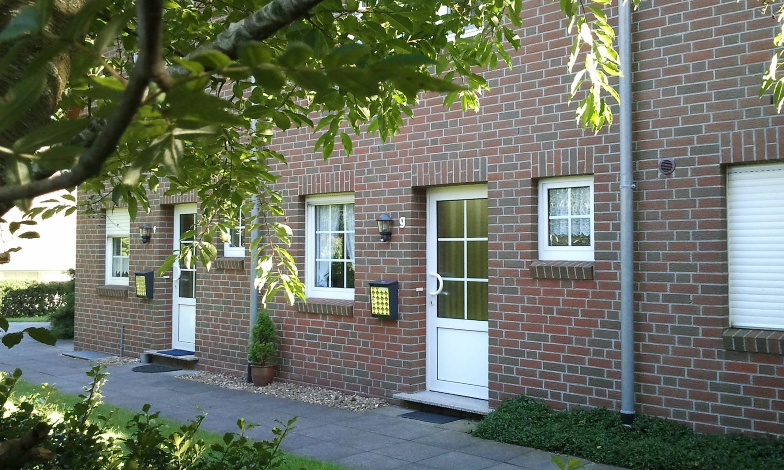 A brick building with multiple windows and doors, a small tree in the foreground, and a paved driveway.