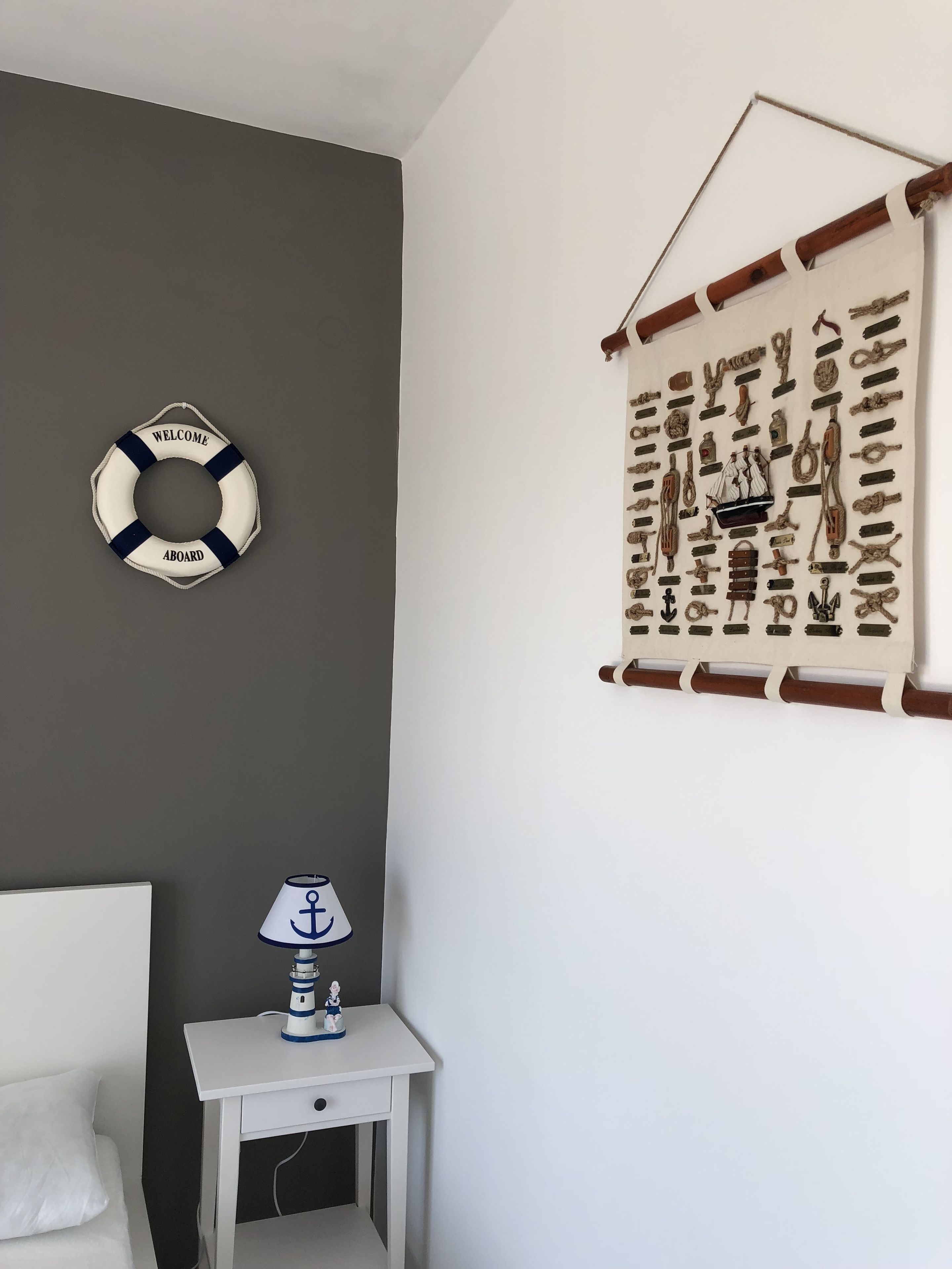 A bedroom with a bed, a nightstand with a lamp, a life preserver, and a nautical-themed wall hanging.