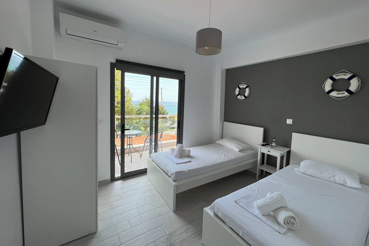 A hotel room with two beds, a TV, a ceiling fan, and a balcony with a view of the sea.