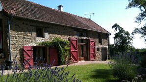 Une ferme en pierre avec des volets rouges, une cheminée en pierre et un jardin où poussent des plants de lavande.