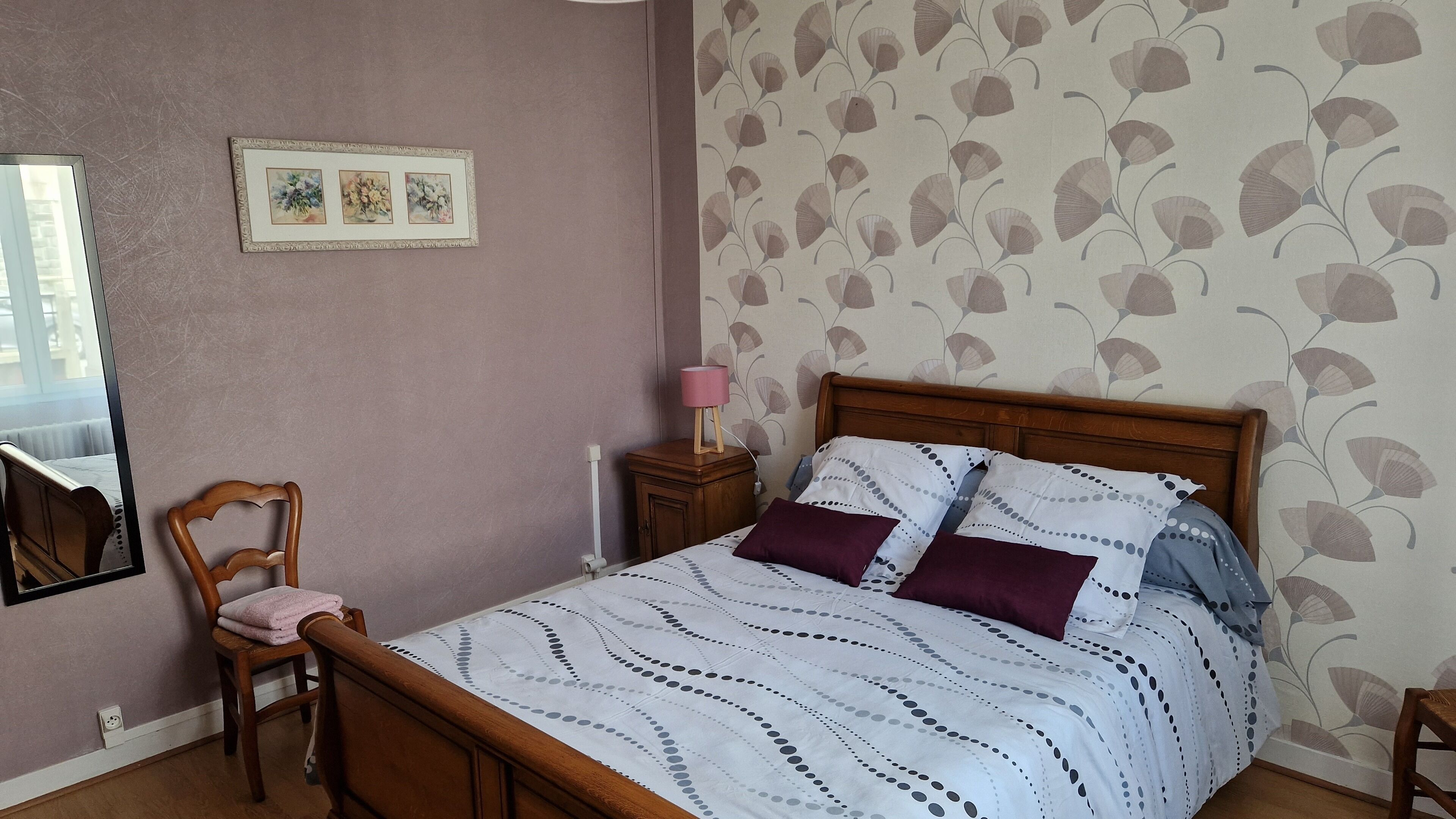 Une chambre avec un lit en bois, une table de chevet avec une lampe, une chaise et un miroir fixé au mur.