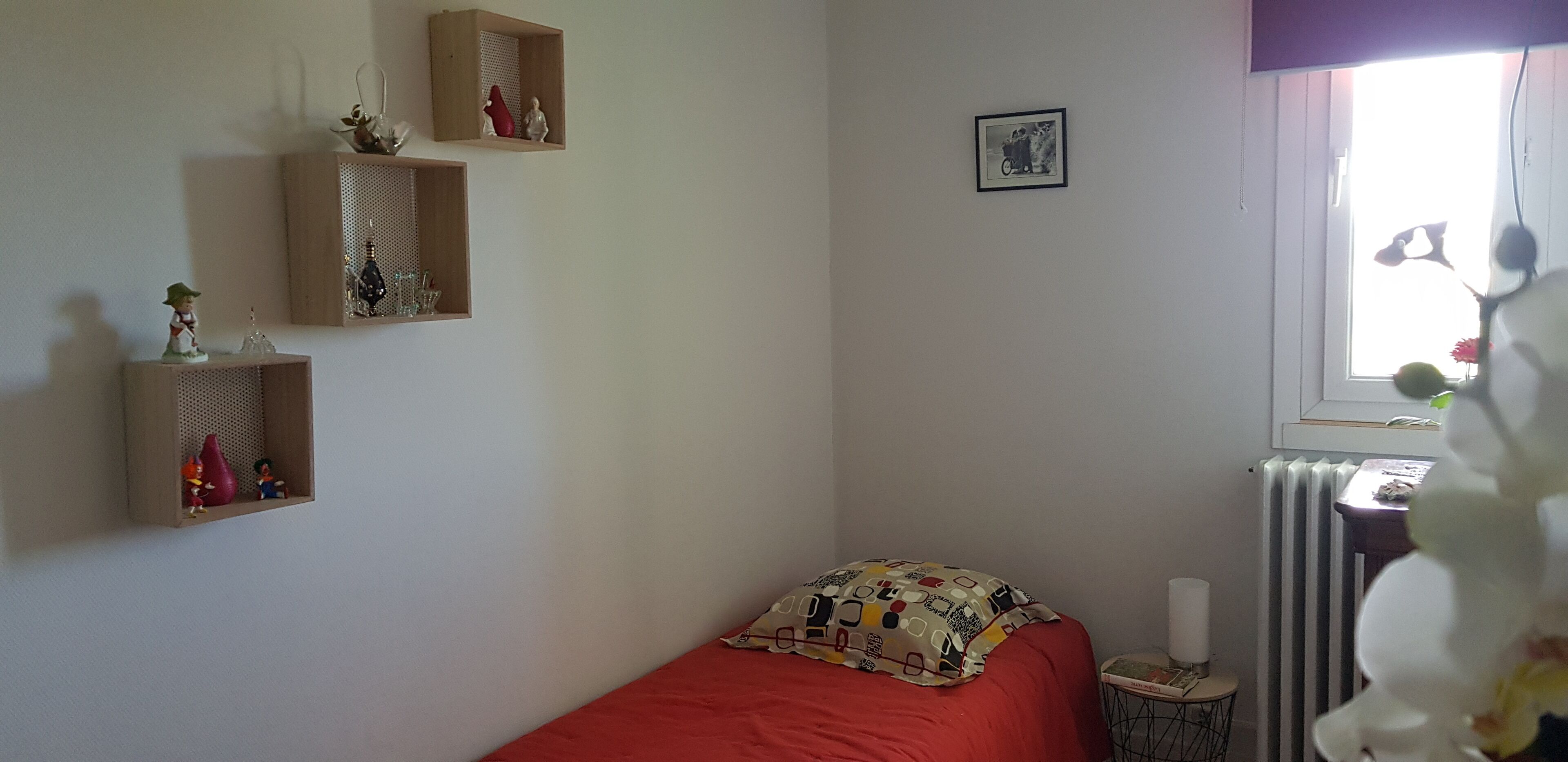 Une chambre avec un lit simple, un radiateur, une petite table avec une lampe et des étagères murales ornées de figurines.
